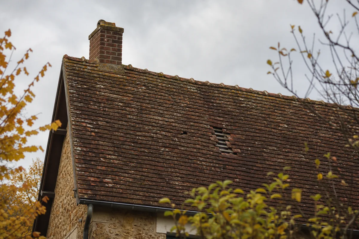 Versant de tuiles plates anciennes sur une maison meulière francilienne, quelques tuiles manquantes au milieu du rang