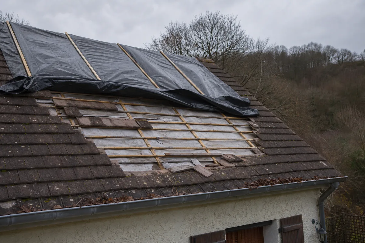 Toiture pavillonnaire en tuile plate partiellement découverte après une tempête, bâche tendue sur le versant, ciel gris d&rsquo;Île-de-France